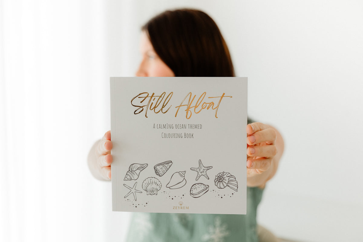 Woman holding up the Still Afloat grief colouring book, showing the gold foil cover and hand drawn shell illustrations